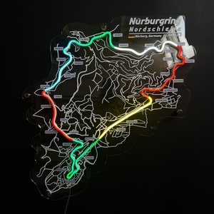 May include: A black and white map of the Nurburgring Nordschleife race track with a neon green, yellow, and red outline. The map is on a black background. The text "Nurburgring Nordschleife" is at the top of the map. The text "Nurburg, Germany 20.832 km" is below the title.