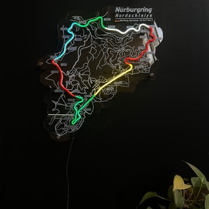 May include: A neon sign depicting the N&uuml;rburgring Nordschleife race track in Germany. The track is outlined in green, yellow, red, and white neon lights. The sign is mounted on a black background.