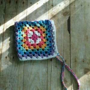 May include: A colorful crocheted granny square with a white border and a long fringe. The square is made with a variety of colors, including blue, green, yellow, orange, pink, and purple.
