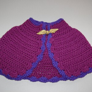 May include: A deep purple crochet capelet featuring a vibrant violet scalloped border along the edges. The garment is secured at the neckline with a metallic gold-toned clasp shaped like two symmetrical wings meeting in the center.