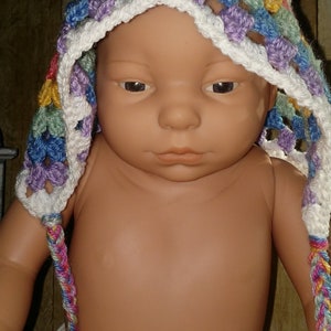 Pu&ograve; includere: Un bambino che indossa un cappello all'uncinetto colorato con un bordo bianco e lunghi lacci. Il cappello &egrave; realizzato con una variet&agrave; di colori, tra cui viola, blu, verde, giallo e bianco.