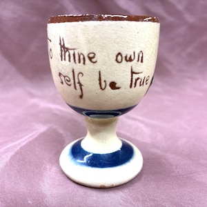 Może przedstawiać: Vintage ceramiczny kieliszek do jajek z kremową podstawą i brązową obwódką. Na kubku widnieje napis "To thine own self be true" w brązowym skrypcie. Podstawa ma niebieskie paski, dodając dekoracyjnego akcentu.