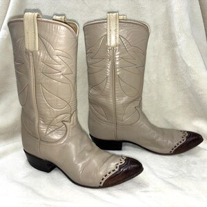 Op de afbeelding: Een paar beige leren cowboylaarzen met een puntige neus en een donkerbruine, getextureerde kap, decoratieve stiksels op de schacht, treklussen aan de bovenkant en een blokhak. De laarzen hebben een gladde afwerking en een western silhouet.