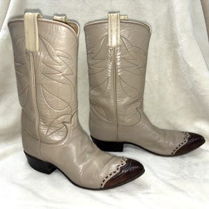 Könnte beinhalten: Ein Paar beigefarbene Cowboystiefel aus Leder mit spitzer Zehenpartie und einer dunkelbraunen, strukturierten Kappe, Ziernähten am Schaft, Zuglaschen am oberen Rand und einem schwarzen Absatz. Die Stiefel haben eine glatte Oberfläche.