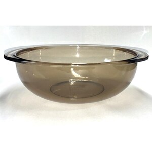 May include: A round, translucent brown glass mixing bowl with a smooth, curved interior and a wide, flat rim that extends outward on two sides to form integrated handles. The glass has a polished finish and a consistent, deep amber tint throughout.
