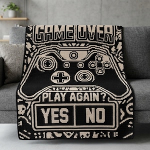May include: A black and cream-coloured throw blanket featuring a pixelated video game controller graphic. The text GAME OVER appears at the top, with PLAY AGAIN? and the options YES and NO displayed in a rectangular box at the bottom of the design.