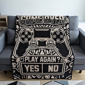 May include: A black and cream-colored crocheted blanket featuring a large video game controller graphic. The text GAME OVER is at the top, with PLAY AGAIN? and the options YES and NO printed in a pixelated font at the bottom of the design.