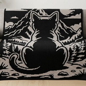 May include: A black and cream-coloured throw blanket featuring a large, centred silhouette of a cat viewed from behind. The cat is positioned in the foreground, looking out towards a detailed landscape of jagged mountains, pine trees, and clouds.