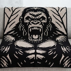 Op de afbeelding: Een rechthoekige gebreide deken met een grote, contrastrijke afbeelding van een brullende gorilla. Het ontwerp gebruikt zwart en beige garen om het gezicht, de gespierde borst en de armen van de gorilla weer te geven, omlijst door gestileerde bladeren.