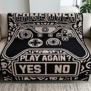 以下が含まれることがあります： 黒とベージュのニットブランケットで、大きなビデオゲームコントローラーのグラフィックが特徴です。上部には「GAME OVER」の文字があり、下部にはピクセル化されたレトロなフォントで「PLAY AGAIN?」というテキストと「YES」「NO」の選択肢が表示されています。