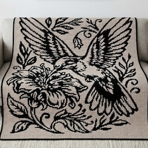 Può includere: Una coperta rettangolare tessuta con sfondo beige e un grande design grafico nero ad alto contrasto raffigurante un colibrì che vola sopra un fiore in piena fioritura, circondato da motivi botanici a foglie e un bordo nero solido.