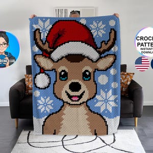 May include: A blue and white crochet blanket featuring a reindeer wearing a Santa hat. The reindeer has brown fur, antlers, and a smiling face. White snowflakes are scattered across the blue background. The blanket is held up by a person.