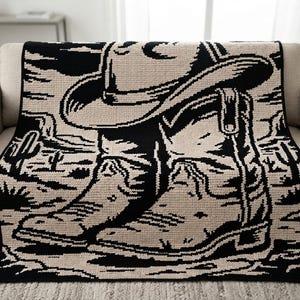 May include: A rectangular knit throw blanket featuring a high-contrast black and cream graphic design. The pattern depicts a cowboy hat resting on a pair of western-style boots, set against a desert landscape background with cacti and mesas.