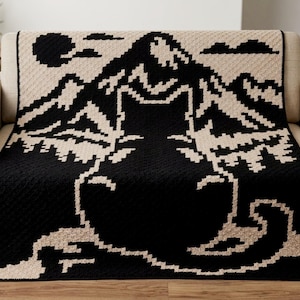 Puede incluir: Una manta de ganchillo rectangular con un diseño pixelado de alto contraste en negro y beige. El patrón muestra un gato grande sentado en primer plano con una cordillera, un sol y nubes al fondo. La textura es uniforme en toda la pieza.
