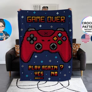 May include: A navy blue crochet blanket with a red pixelated video game controller design. The blanket features the words "GAME OVER" and "PLAY AGAIN? YES NO" in a pixelated font. The blanket is displayed against a neutral background.