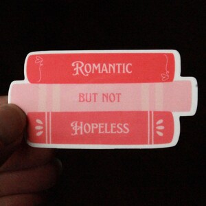May include: A sticker featuring three stacked book-shaped designs. The top book is red with the word "ROMANTIC" in white. The middle book is light pink with the words "BUT NOT" in white. The bottom book is red with the word "HOPELESS" in white.