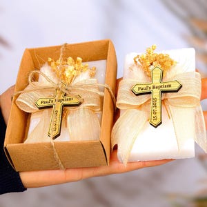Könnte beinhalten: Zwei kleine weiße Seifen-Geschenke mit einem Holzkreuz, das an jedem befestigt ist. Das Kreuz ist mit "Paul's Baptism" und dem Datum "25.07.2025" graviert. Jede Seife ist in ein beigefarbenes Band gewickelt und mit getrockneten Blumen verziert.