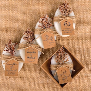Peut inclure: Savons blancs ovales décorés de fleurs séchées et de ficelle, chacun avec une étiquette personnalisée. Les étiquettes affichent des noms et des dates de mariage. Un savon est dans une petite boîte carrée. Le fond est un tissu texturé beige.