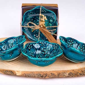 Puede incluir: Cuatro cuencos de cerámica turquesa con diseño floral, presentados sobre una tabla de madera. Un juego está empaquetado en una caja con una etiqueta "Bober & Michael". Los cuencos tienen una superficie texturizada y un borde festoneado.