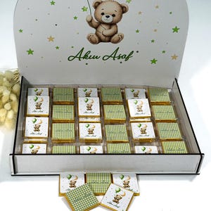 Op de afbeelding: Een witte rechthoekige doos met vierentwintig vierkante chocolaatjes in goudfolie met groen ruitpatroon of teddybeer-afbeeldingen. Het deksel toont een illustratie van een teddybeer en de tekst Aku Asaf met groene en gouden sterren.