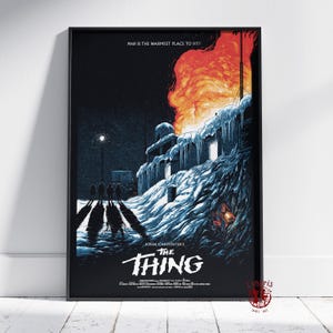 May include: A framed movie poster for John Carpenter's "The Thing." The artwork depicts a snow-covered building engulfed in flames, with silhouetted figures in the foreground. The text "MAN IS THE WARMEST PLACE TO HIDE!" is at the top.