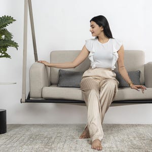 Può includere: Una donna è seduta su un divano sospeso beige con un cuscino a quadri bianco e nero. Il divano è sospeso dal soffitto con corde spesse. La donna indossa una camicetta bianca con volant e pantaloni beige.