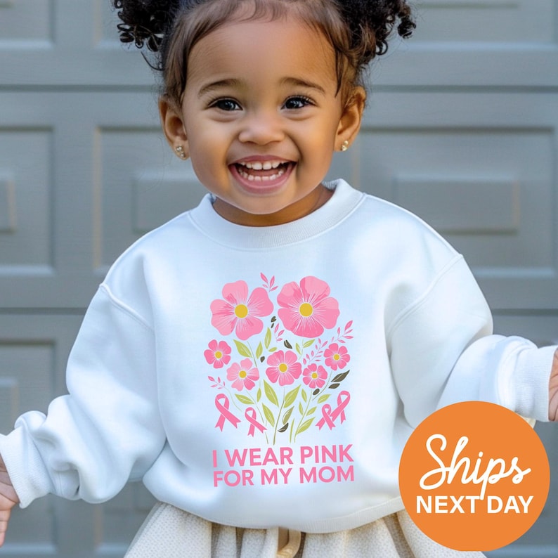 A young girl with curly hair is smiling and wearing a white sweatshirt with a pink flower design and the text "I WEAR PINK FOR MY MOM".