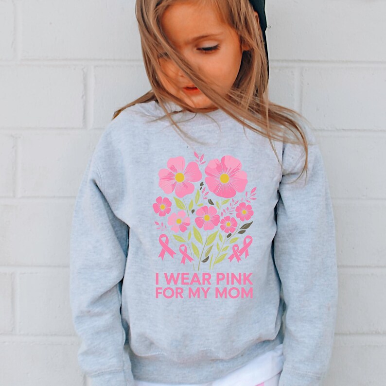 A young girl wearing a gray sweatshirt with a pink floral design and the text "I WEAR PINK FOR MY MOM" printed on it.