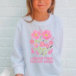 A young girl wearing a white sweatshirt with a pink flower design and the text "I WEAR PINK FOR MY MOM" printed on it.