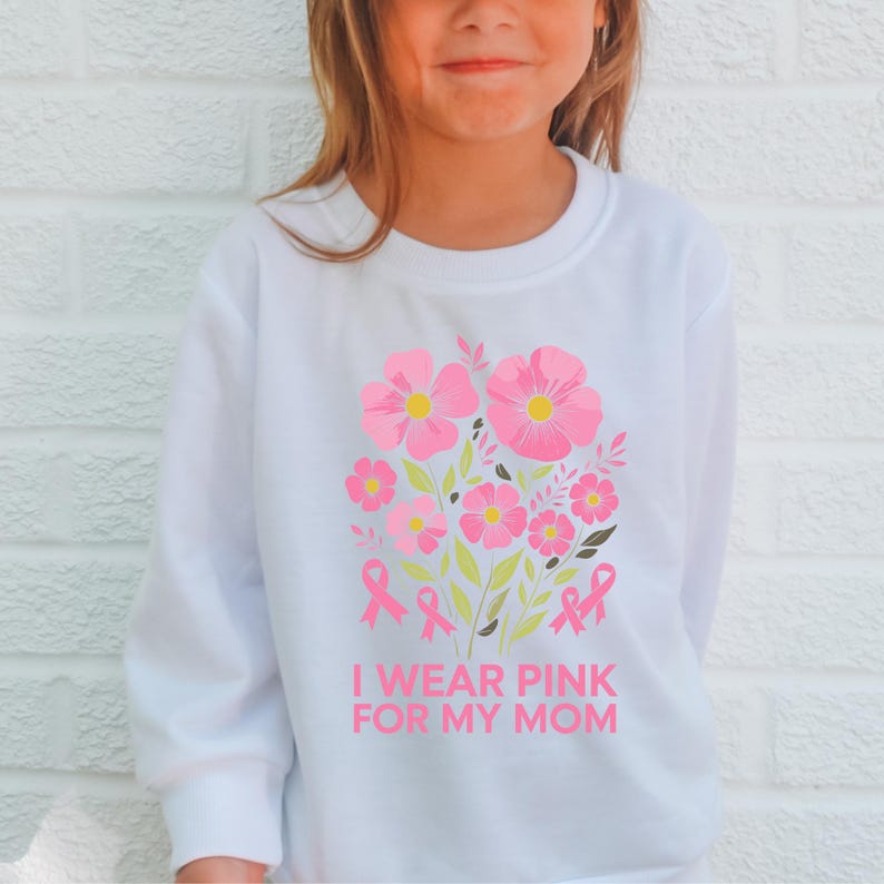A young girl wearing a white sweatshirt with a pink flower design and the text "I WEAR PINK FOR MY MOM" printed on it.