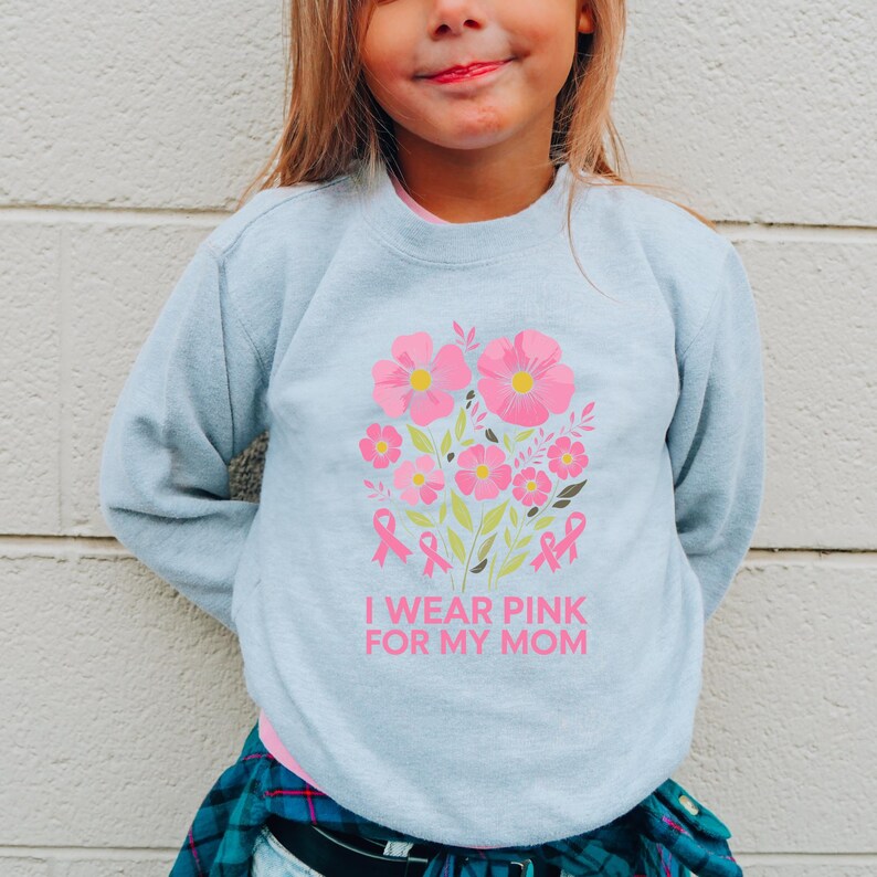 A young girl wearing a light blue sweatshirt with a pink floral design and the text "I WEAR PINK FOR MY MOM" printed on it.
