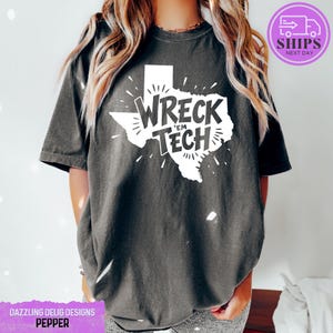 May include: A dark gray t-shirt with a white outline of the state of Texas. Inside the outline is the text "Wreck 'Em Tech" in a white, handwritten font.