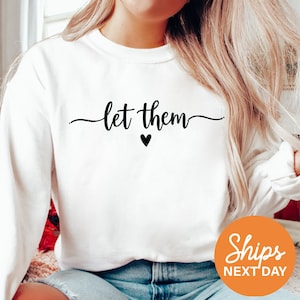 May include: White sweatshirt with the text "let them" in a black script font, with a small black heart below the text. The sweatshirt has long sleeves and a crew neck. An orange circle with the words "Ships Next Day" is in the lower right corner.