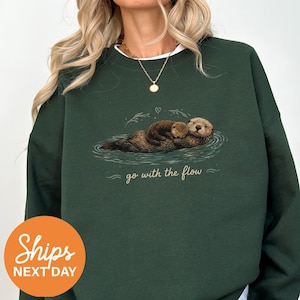 May include: A dark forest green crewneck sweatshirt featuring a graphic illustration of two brown sea otters floating in water. Below the otters, the text "go with the flow" is printed in a light beige script font, accompanied by a small heart.