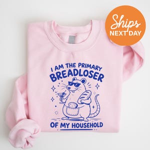 May include: A light pink crewneck sweatshirt featuring a blue graphic of a rat wearing sunglasses, holding a martini glass and a loaf of bread, with the text I AM THE PRIMARY BREADLOSER OF MY HOUSEHOLD printed in blue lettering above and below the rat.
