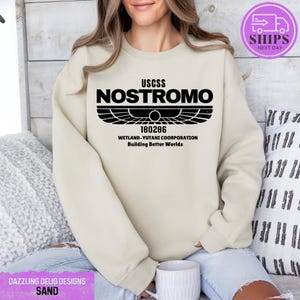 May include: A beige sweatshirt with a black graphic print of the text "USCSS NOSTROMO" with a winged symbol and the text "180286 WETLAND-YUTANI COORPORATION Building Better Worlds".