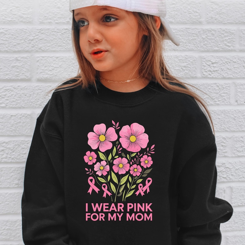 A young woman wearing a black sweatshirt with a pink flower design and the text "I WEAR PINK FOR MY MOM" printed on it.
