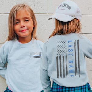 May include: A light blue sweatshirt with a small American flag graphic on the front and a larger distressed flag design on the back. The back design includes the text "LAST NAME" in blue. A white baseball cap is also visible.