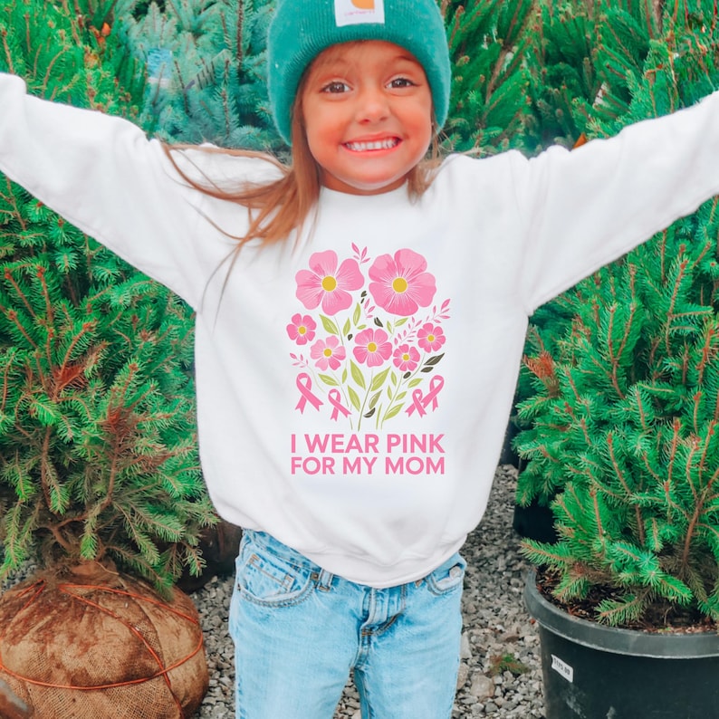 A young girl wearing a white sweatshirt with pink flowers and pink ribbons stands in front of a group of potted plants.