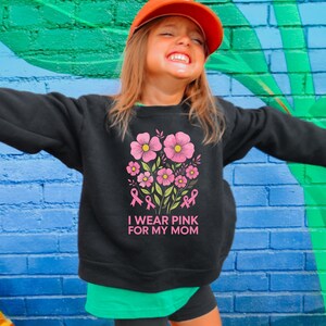 A young girl wearing a black sweatshirt with a pink flower design and the text "I WEAR PINK FOR MY MOM" smiling and looking up.