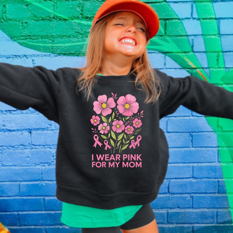 A young girl wearing a black sweatshirt with a pink flower design and the text "I WEAR PINK FOR MY MOM" smiling and looking up.