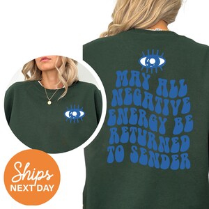 May include: Dark green sweatshirt with a blue eye graphic on the front and back. The back of the sweatshirt has the text "MAY ALL NEGATIVE ENERGY BE RETURNED TO SENDER" in blue. The sweatshirt is a crew neck style.
