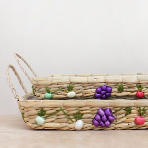 May include: Two rectangular woven baskets with handles. The baskets are decorated with colorful, embroidered fruit and vegetable designs, including grapes, radishes, and tomatoes. The baskets are made of natural-colored woven material.