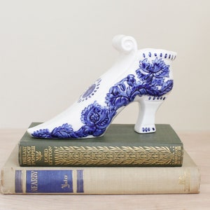 May include: A white ceramic shoe-shaped vase with blue floral designs. The heel is high and the shoe is resting on two vintage books with gold lettering. The vase is a decorative item.