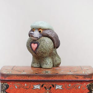 May include: A ceramic poodle figurine with a heart-shaped design on its chest, perched atop a decorative orange tin. The poodle has a green body, brown fur, and a light green hat. The tin features ornate gold and black patterns.