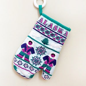 May include: A teal blue oven mitt with a white background and pink and purple accents. The mitt features a traditional Alaskan design with two figures in parkas and the word "ALASKA" in pink letters.