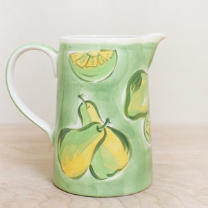 May include: A green ceramic pitcher with a white handle. The pitcher is decorated with a hand-painted design of pears and lemons.