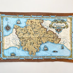 May include: A rectangular textile featuring a vintage map of Devon, England. The map is in shades of tan, blue, and brown, with illustrations of ships and a compass rose. The text "Devon" is displayed at the top.