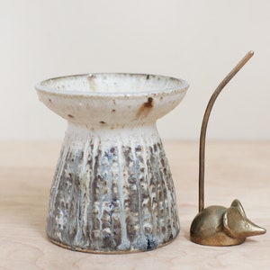 May include: A white ceramic vase with a textured brown and gray glaze. The vase has a wide, flat top and a narrow base. A small gold metal mouse figurine with a long tail is sitting next to the vase.