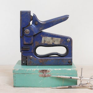 May include: A blue metal staple gun with a black handle, sitting on top of a teal wooden box. A silver compass is lying on the box.